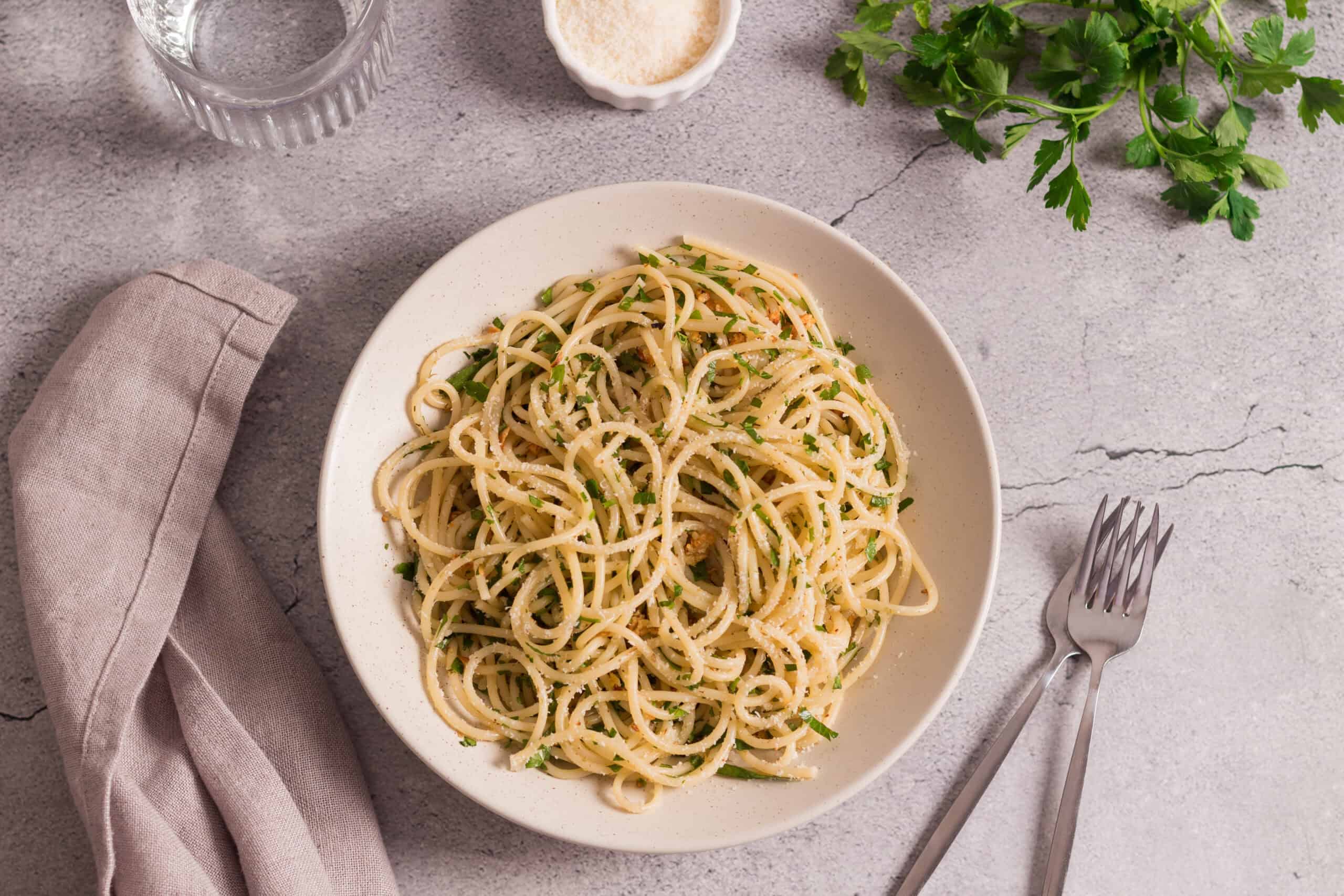 Creamy garlic spaghetti pasta topped with parsley, served on a white plate with a glass of water, salt, and fresh herbs on a textured gray surface. Perfect for quick, delicious Italian-inspired meals.