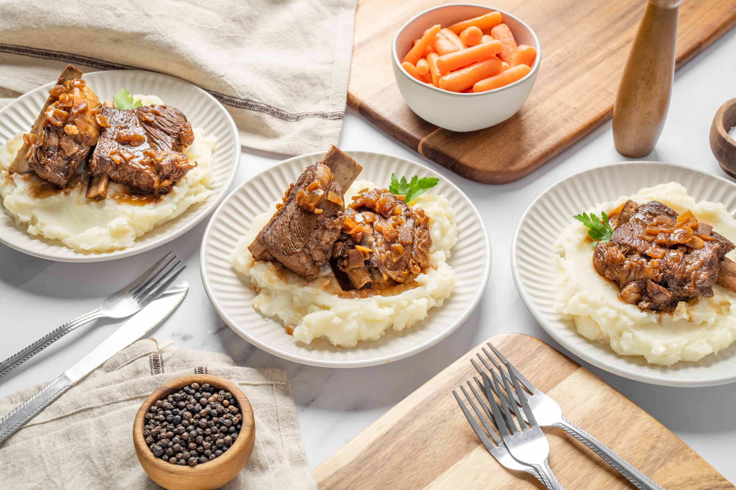 Braised beef short ribs served over creamy mashed potatoes with herb garnish, complemented by a bowl of carrots; hearty comfort food with rich gravy.