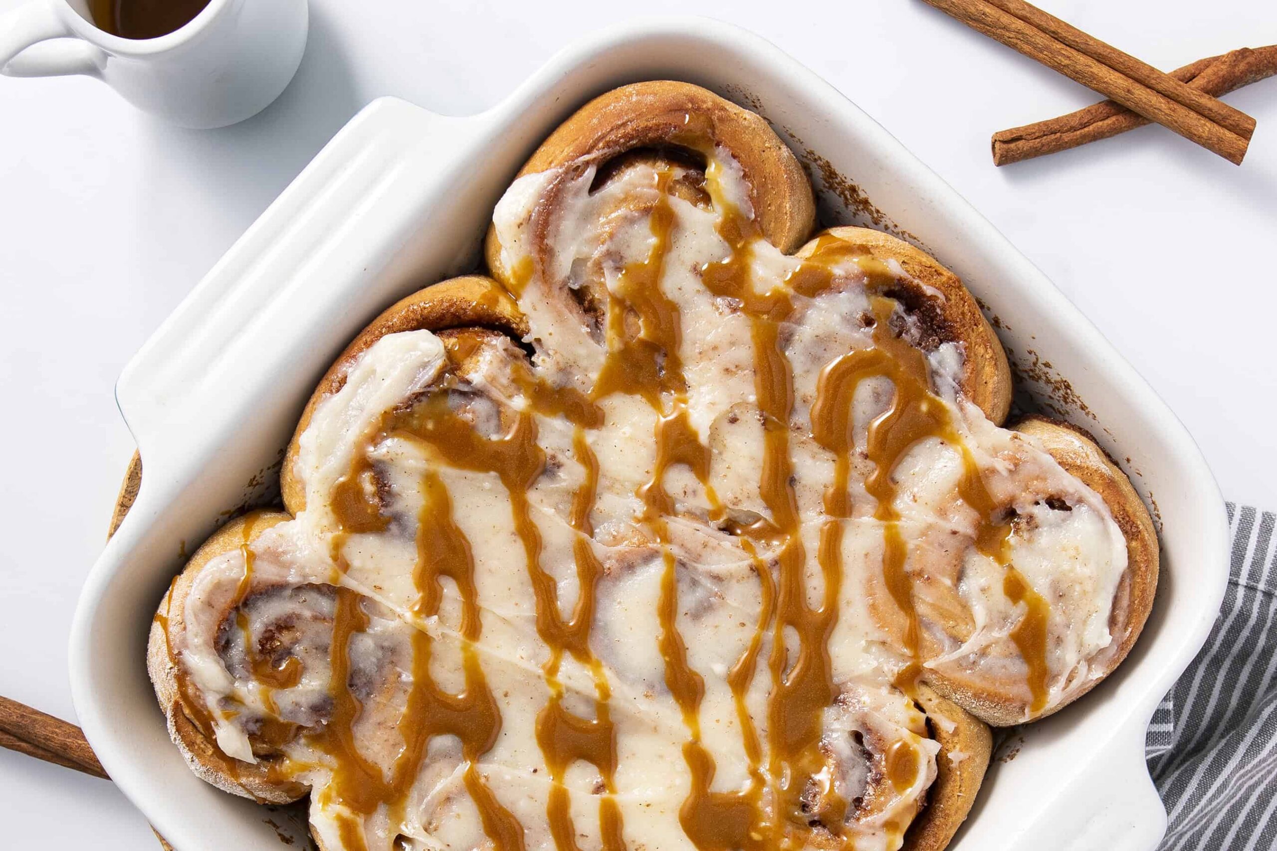 Creamy cinnamon rolls with caramel sauce in a white baking dish, topped with melted icing and drizzled caramel, served as a delicious breakfast or dessert option.