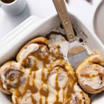 Creamy cinnamon rolls with vanilla glaze and caramel drizzle on a white baking dish, served with extra cinnamon roll pieces on side for a delicious breakfast or dessert.