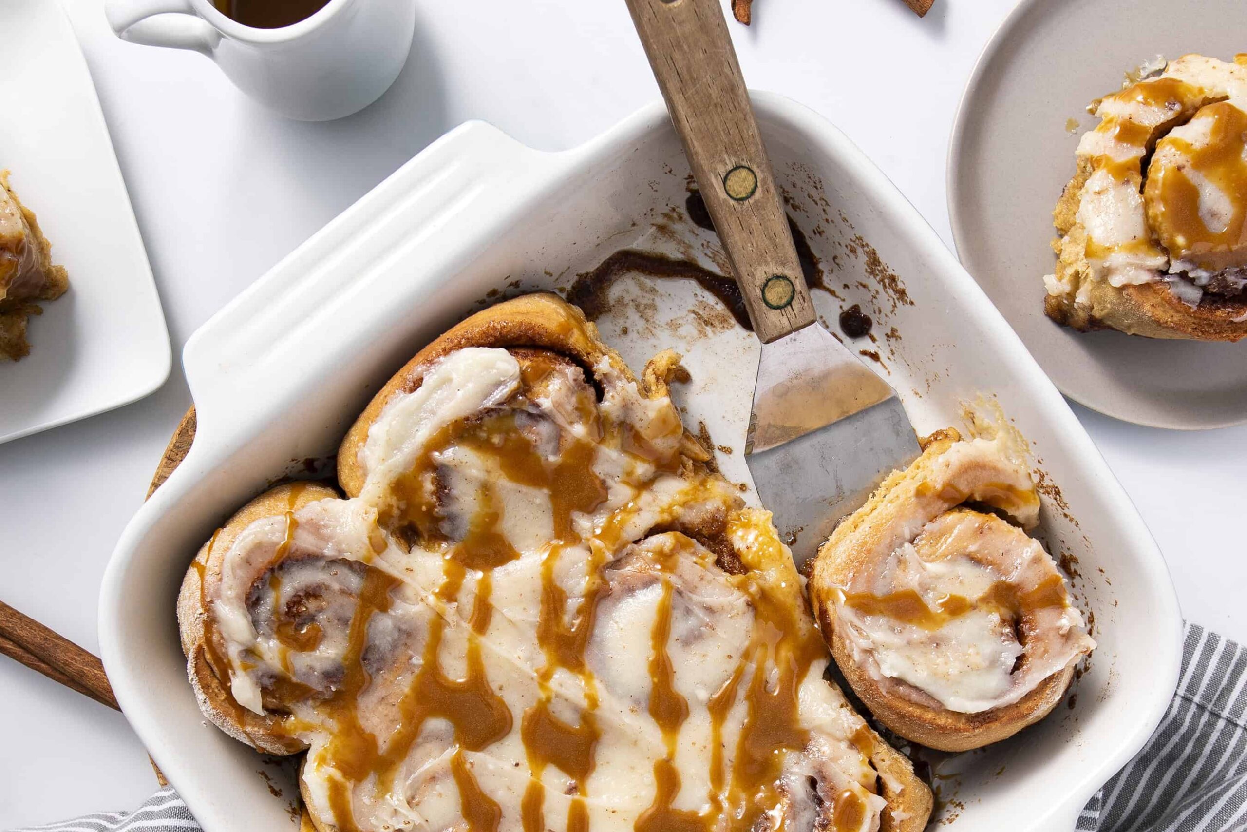 Creamy cinnamon rolls with vanilla glaze and caramel drizzle on a white baking dish, served with extra cinnamon roll pieces on side for a delicious breakfast or dessert.