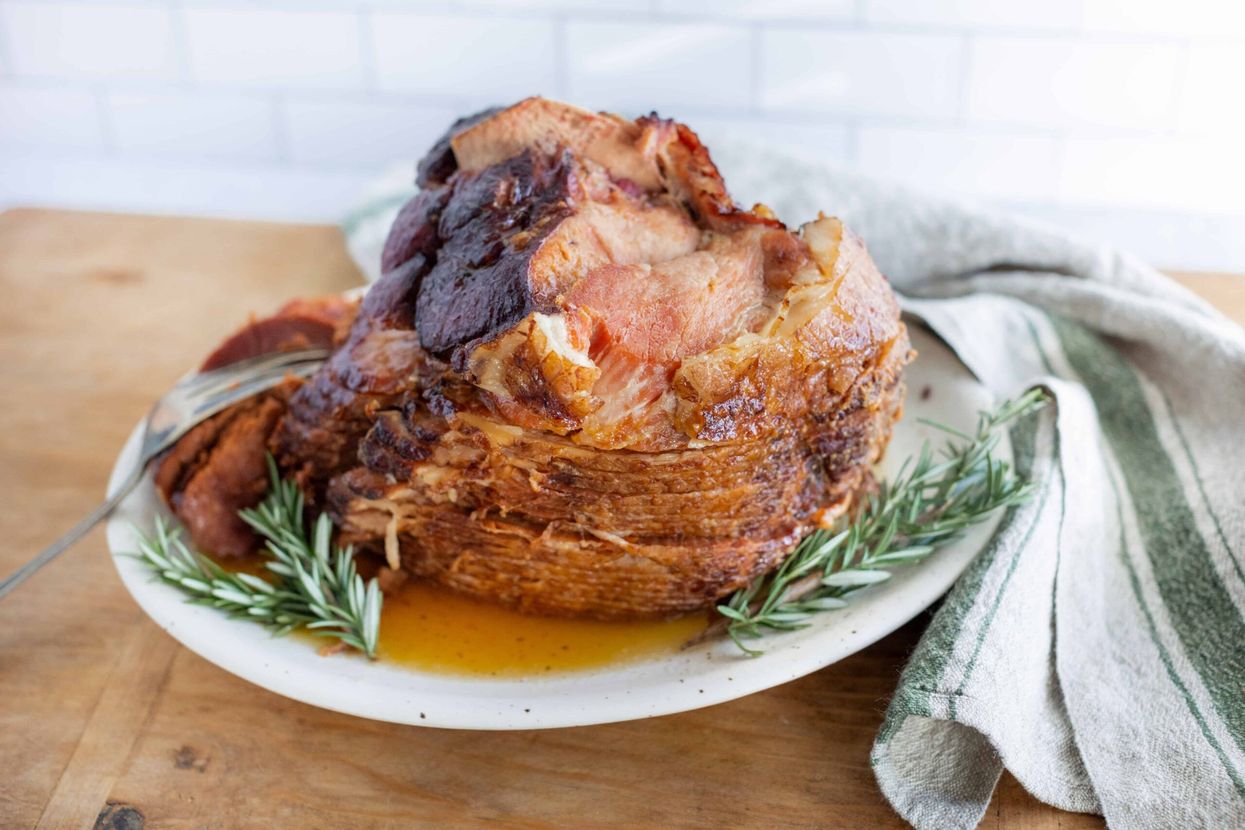 Juicy glazed ham with crispy edges garnished with fresh rosemary on a white plate, served on a rustic wooden table for a delicious holiday meal.