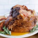 Juicy baked ham with a caramelized glaze garnished with fresh rosemary on a white platter. Perfect for holiday dinner recipes and festive celebrations.
