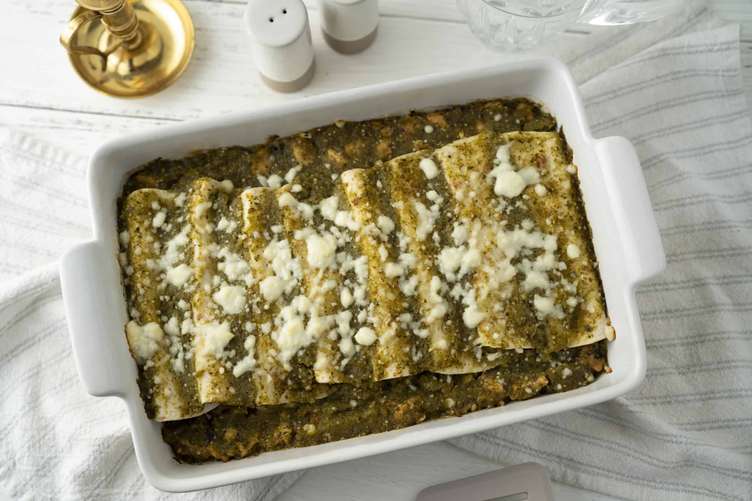 Baked stuffed pasta with rich green sauce, topped with crumbled cheese in a white casserole dish, served on a white wooden table with salt and pepper shakers and glass of water.