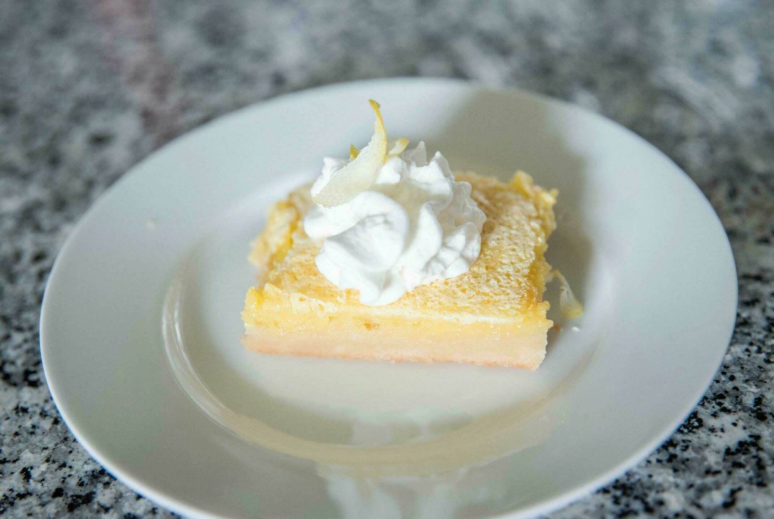 Buttermilk lemon bar with whipped cream on a white plate in front of a gray stone countertop, fresh baked dessert with lemon zest and powdered sugar, perfect for spring or summer treats, from Hey Recipe site.
