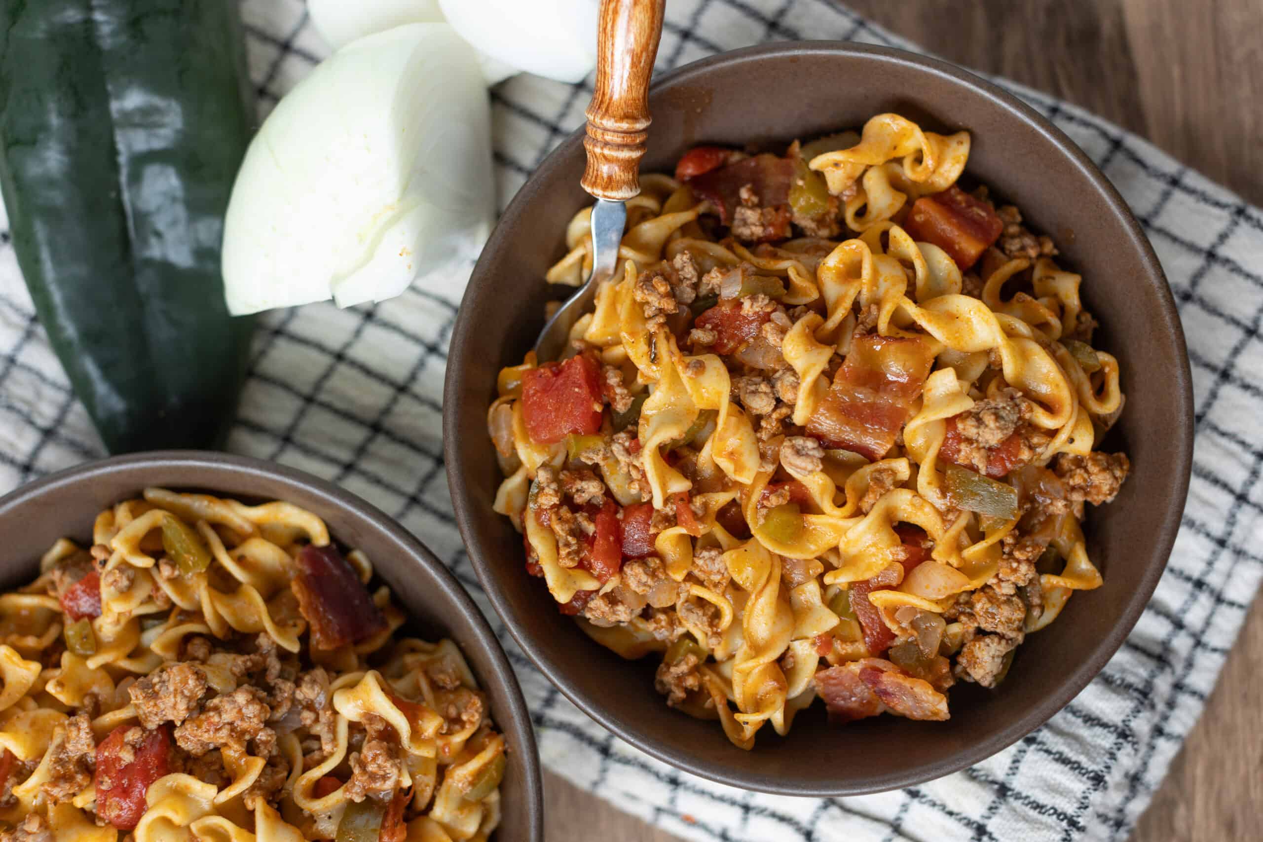 Savory beef and vegetable pasta dish in brown bowls, garnished with fresh zucchini and black pepper on a checkered cloth.