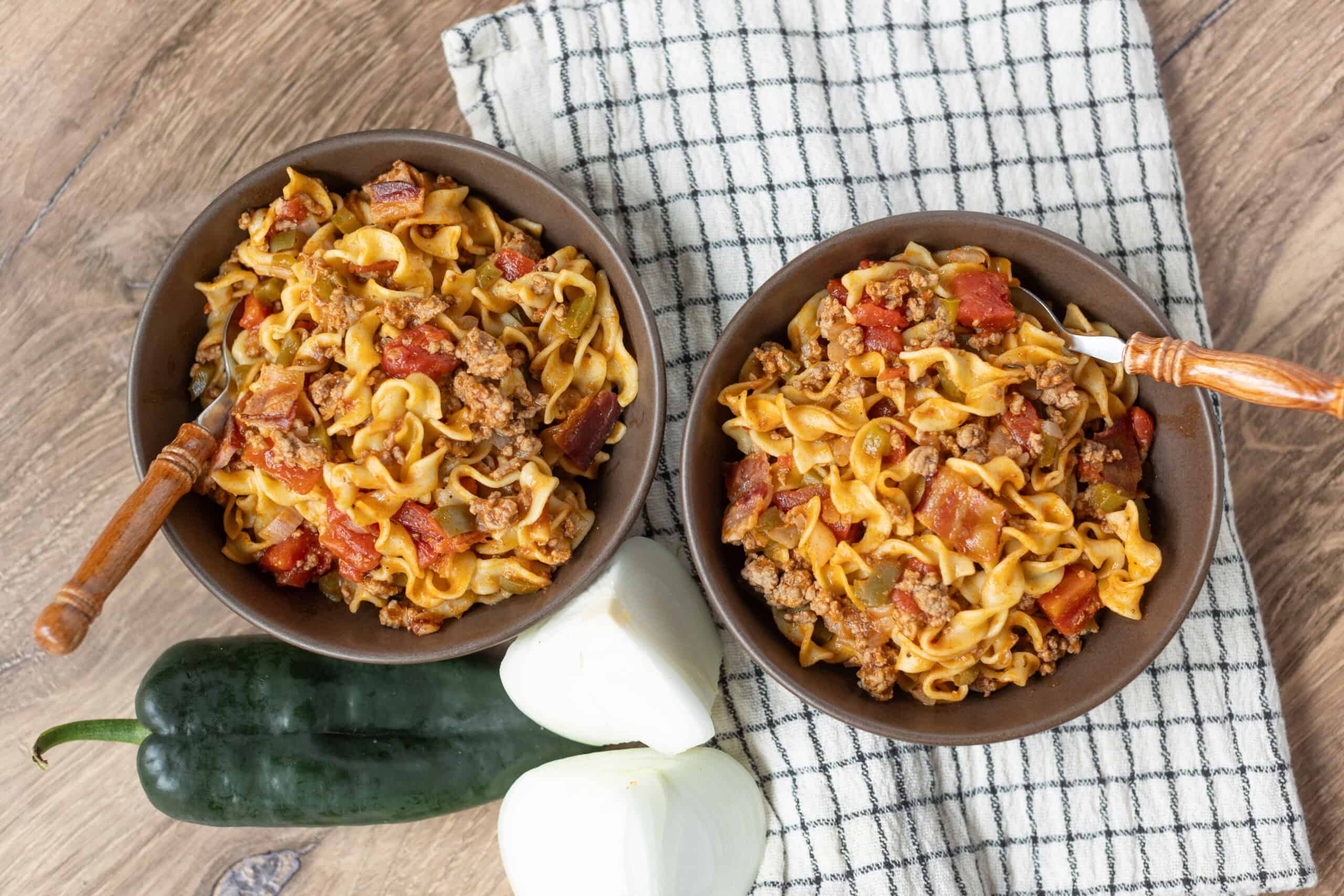 Creamy ground beef and vegetable pasta served in two brown bowls on a rustic wooden table with a checkered cloth, garnished with fresh zucchini and onions, perfect for a comforting homemade meal.