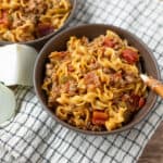 Creamy beef and vegetable noodle stir-fry in two dark bowls with onion wedges and a checkered cloth on a wooden surface, showcasing a hearty and flavorful homemade comfort food recipe.