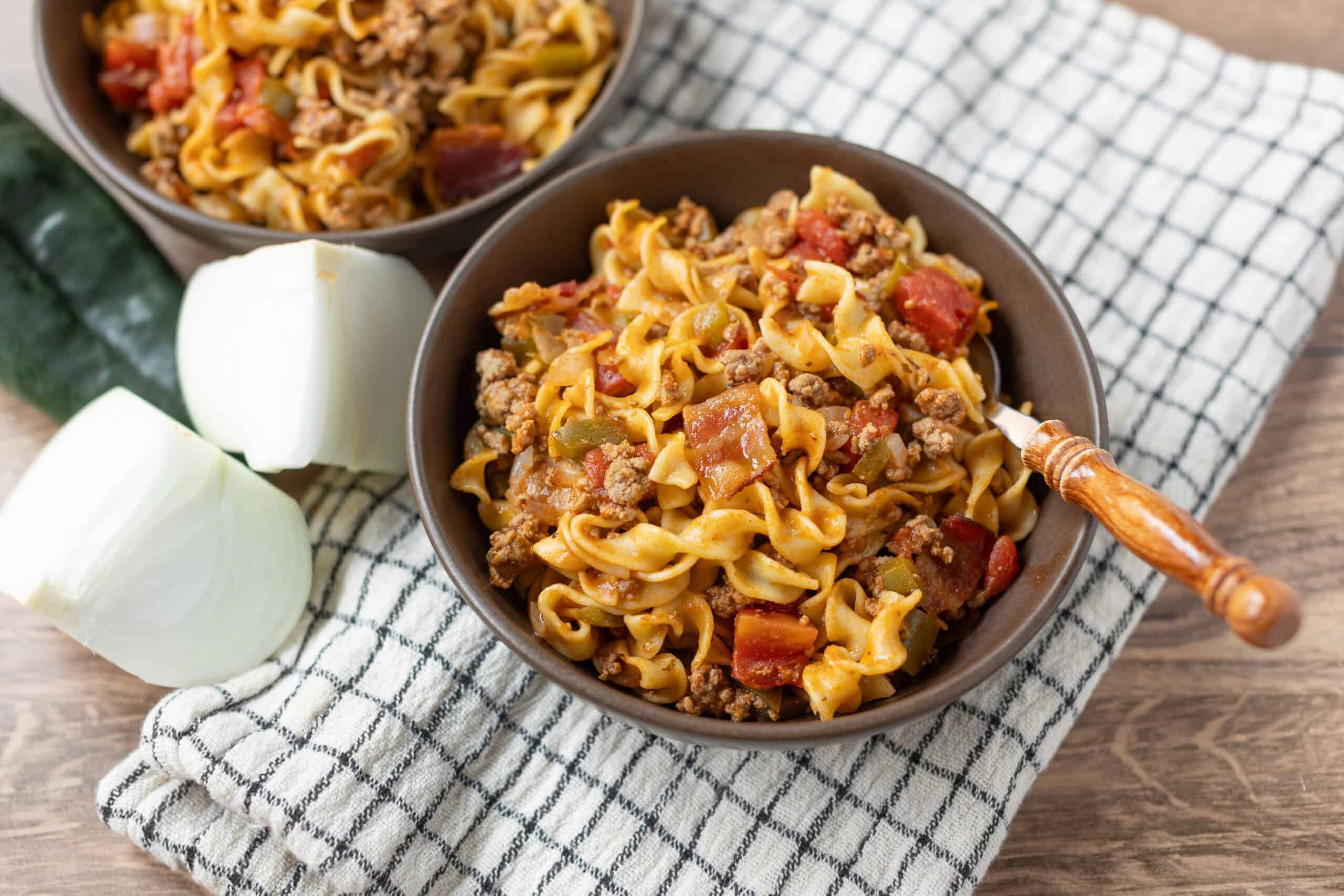 Creamy beef and vegetable noodle stir-fry in two dark bowls with onion wedges and a checkered cloth on a wooden surface, showcasing a hearty and flavorful homemade comfort food recipe.