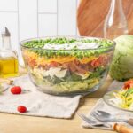 Fresh classic layers of chicken, eggs, cheese, and vegetables in a large glass bowl for a hearty chicken salad recipe. Perfect for lunch or dinner, served with fresh side salads and healthy ingredients.