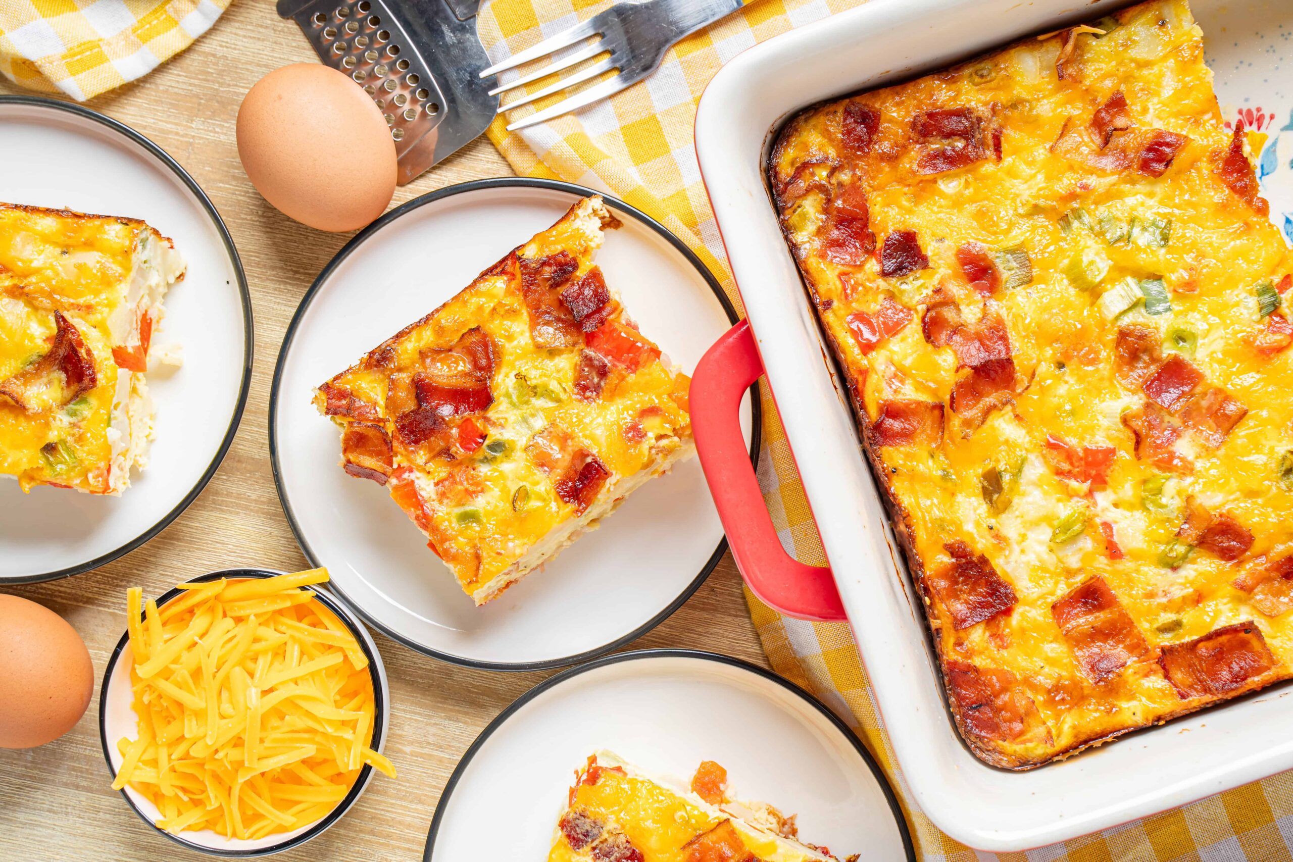 Cheesy baked breakfast casserole with eggs, cheese, and bacon served with fresh eggs and shredded cheese on a wooden table.