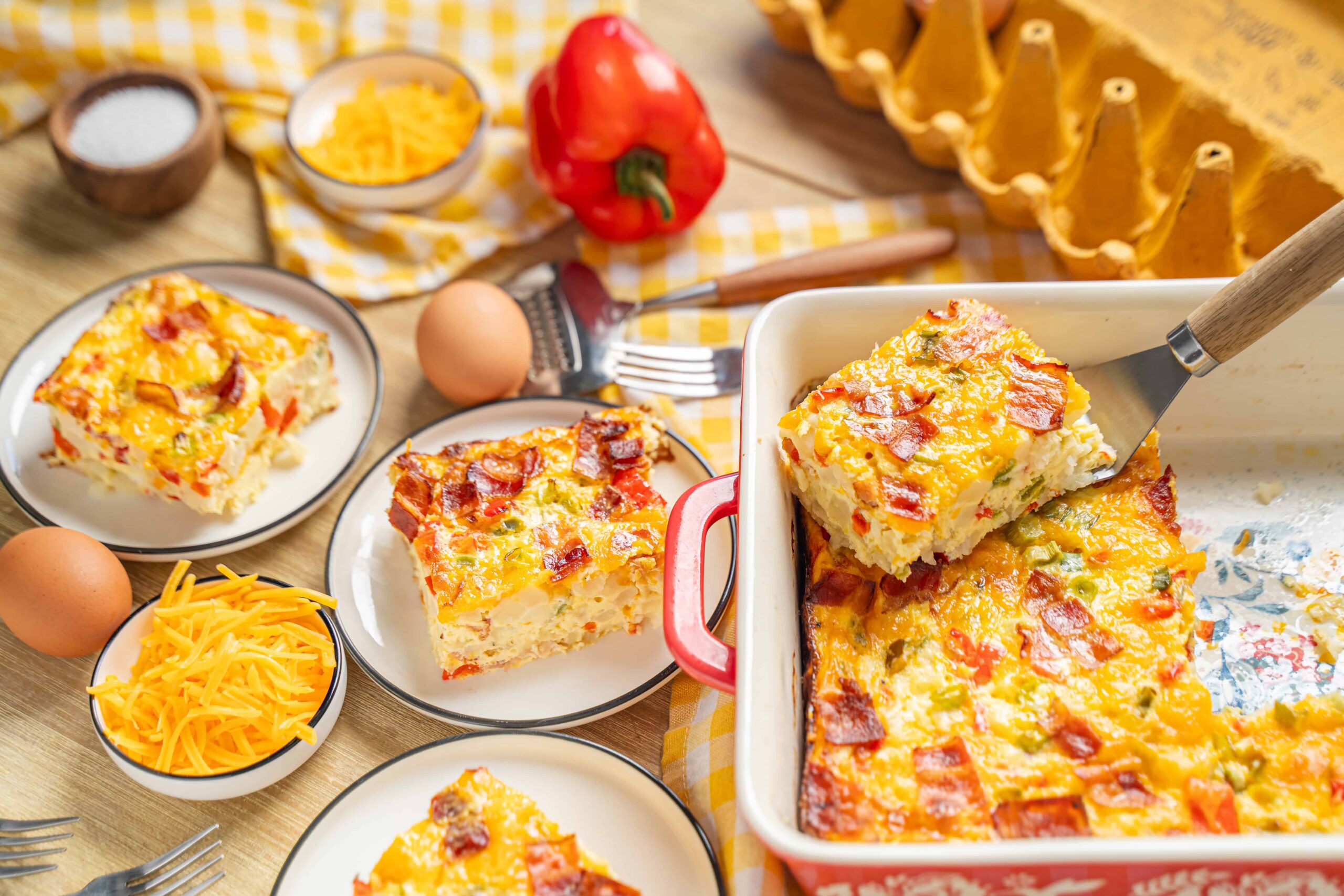 Savory breakfast casserole with cheese, bacon, and vegetables in a baking dish with slices served on plates, surrounded by eggs, shredded cheese, and fresh vegetables on a wooden table.