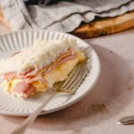 Savory ham and cheese mashed potato casserole on a white plate with a fork, served as a comforting meal. Perfect for family dinners, with fluffy mashed potatoes topped with ham and melted cheese.