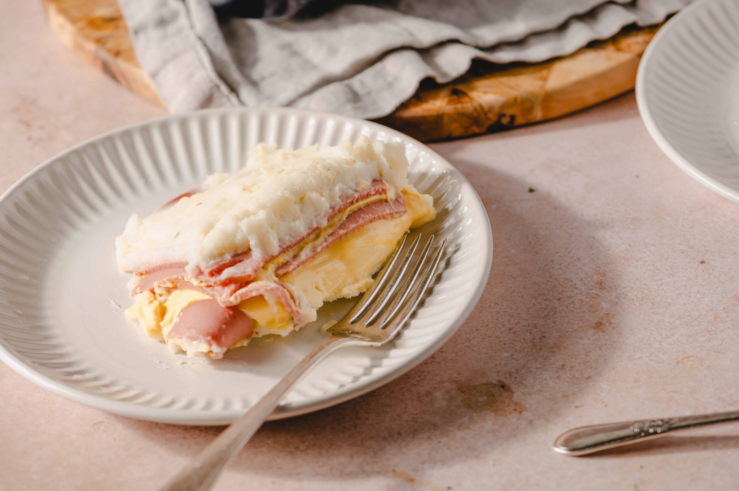 Savory ham and cheese mashed potato casserole on a white plate with a fork, served as a comforting meal. Perfect for family dinners, with fluffy mashed potatoes topped with ham and melted cheese.