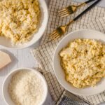 Creamy Parmesan cheese risotto served in white bowls, garnished with grated cheese, with cooking ingredients like cheese block and grated cheese nearby on a white rustic table.