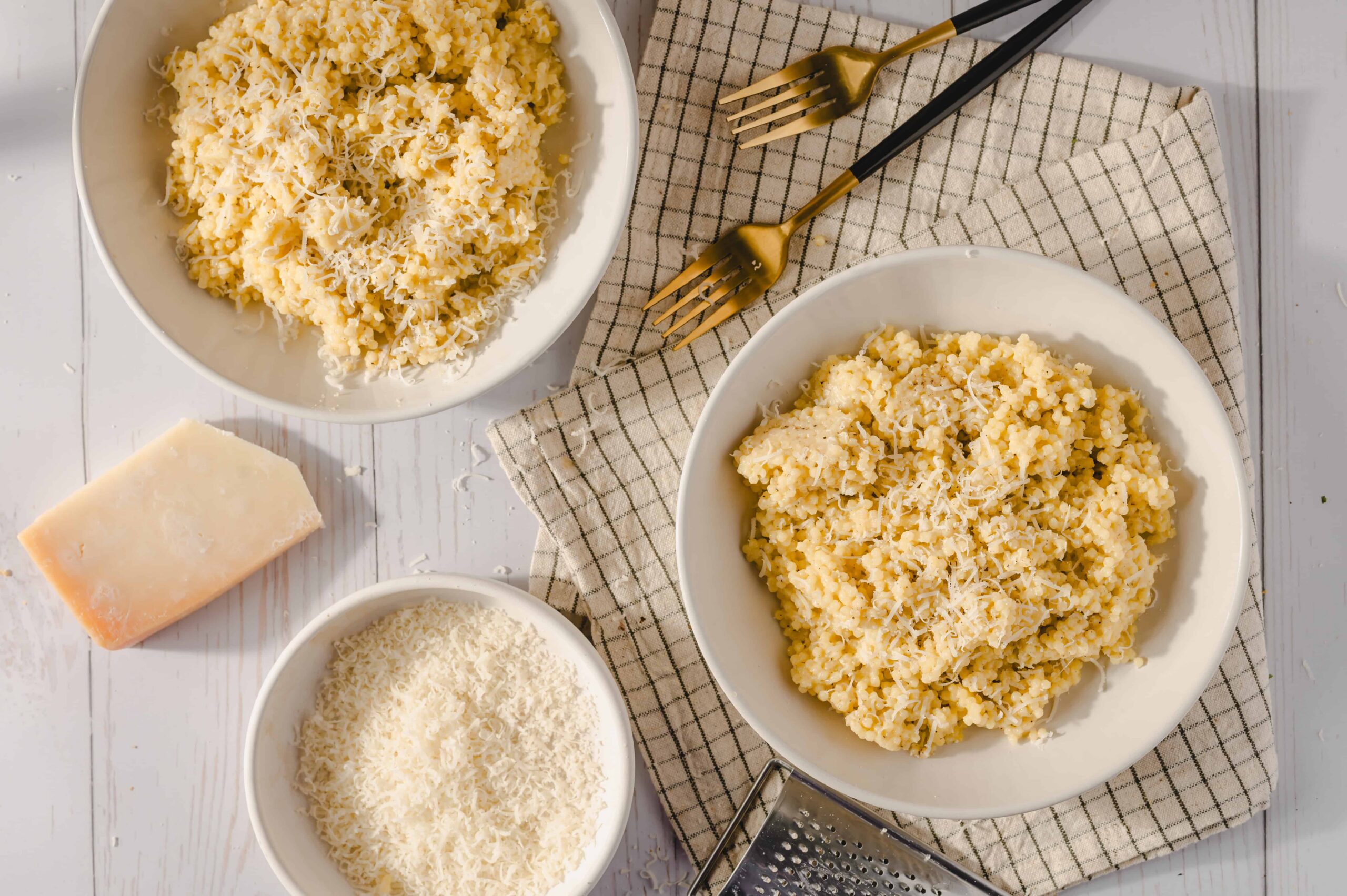 Creamy Parmesan cheese risotto served in white bowls, garnished with grated cheese, with cooking ingredients like cheese block and grated cheese nearby on a white rustic table.