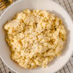 Creamy cheese mashed potatoes with shredded cheese on top, served in a white bowl on a checkered tablecloth. Perfect comfort food for dinner or holiday meals.