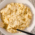 Creamy grated cheese on fluffy mashed potatoes served on a white plate, garnished with shredded cheese, with a gold fork on the side, perfect comfort food.
