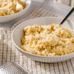 Creamy shredded cheese pasta in a white bowl with grated cheese and additional bowls of cheese nearby, on a rustic kitchen setup for a delicious homemade meal.