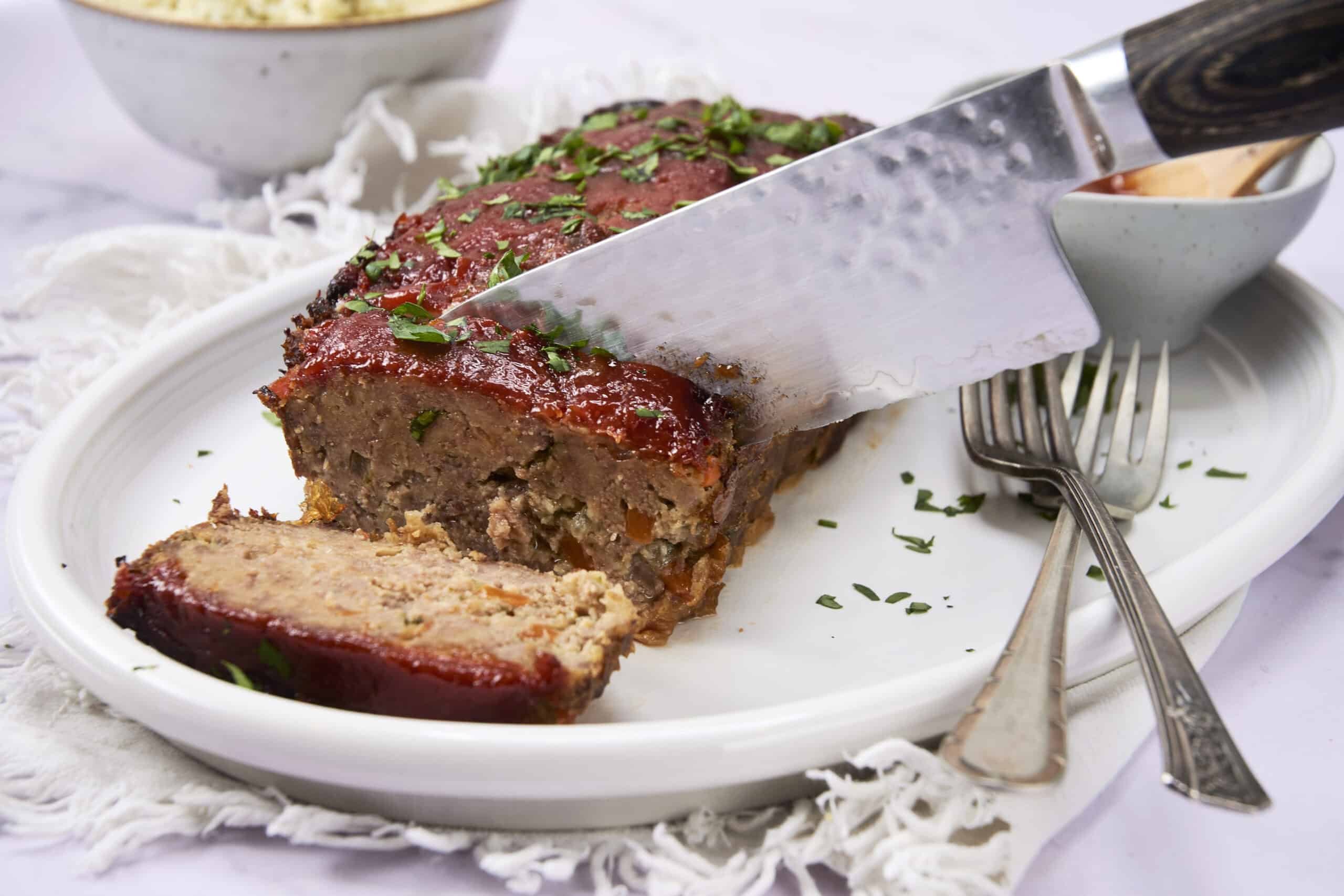 Roast meatloaf with a savory glazed top, garnished with chopped herbs, sliced and served on a white plate with vintage forks, ideal for comfort food recipes.