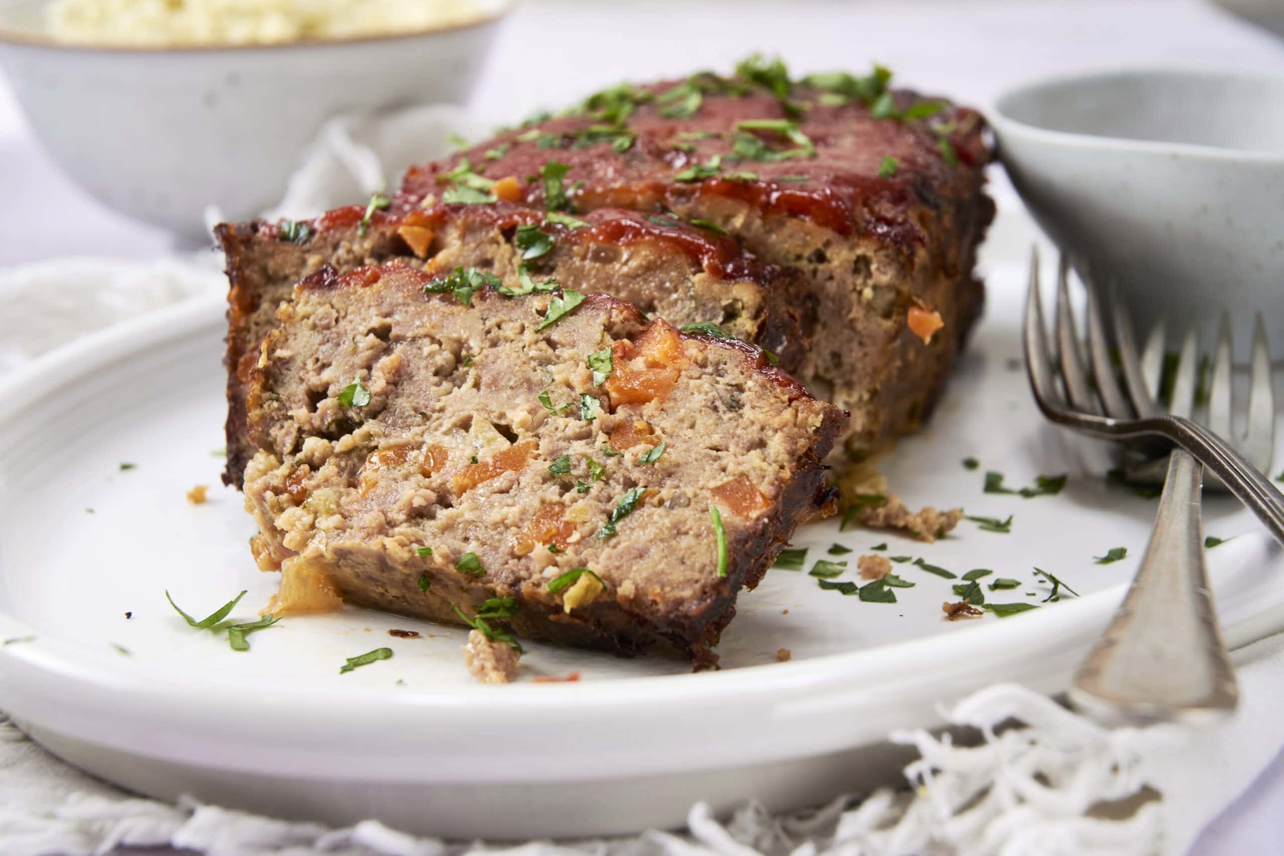 Sighted meatloaf with vegetables sliced on a white plate, garnished with chopped parsley, accompanied by small bowls of sauces or condiments, on a white tablecloth; close-up of a delicious, hearty meatloaf dish.