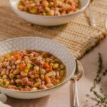 Creamy white bean and ham soup served in a patterned white bowl, garnished with herbs, on a rustic table with a beige cloth and sprigs of thyme, perfect for comforting home-cooked meals.