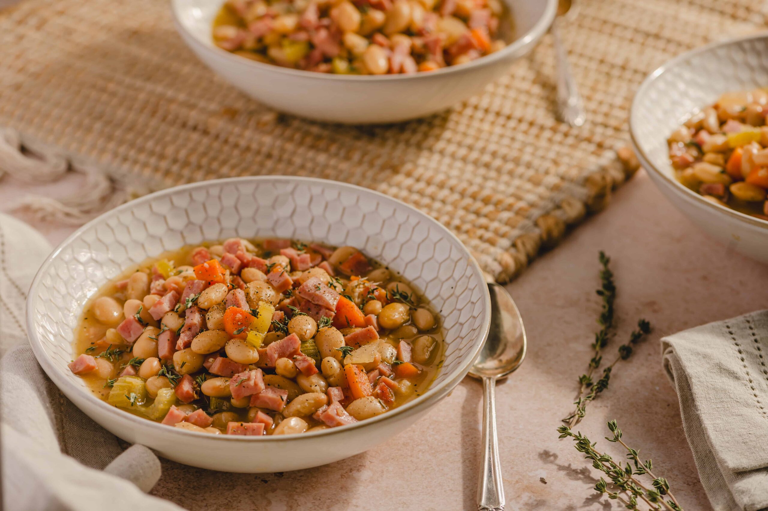 Creamy white bean and ham soup served in a patterned white bowl, garnished with herbs, on a rustic table with a beige cloth and sprigs of thyme, perfect for comforting home-cooked meals.