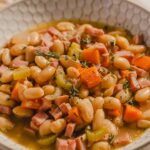 Creamy white beans and ham stew with carrots and herbs in a white textured bowl for hearty comfort food.