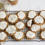 Soft cookies topped with white icing and sprinkles cooling on a wire rack with a bowl of icing and ingredients nearby.