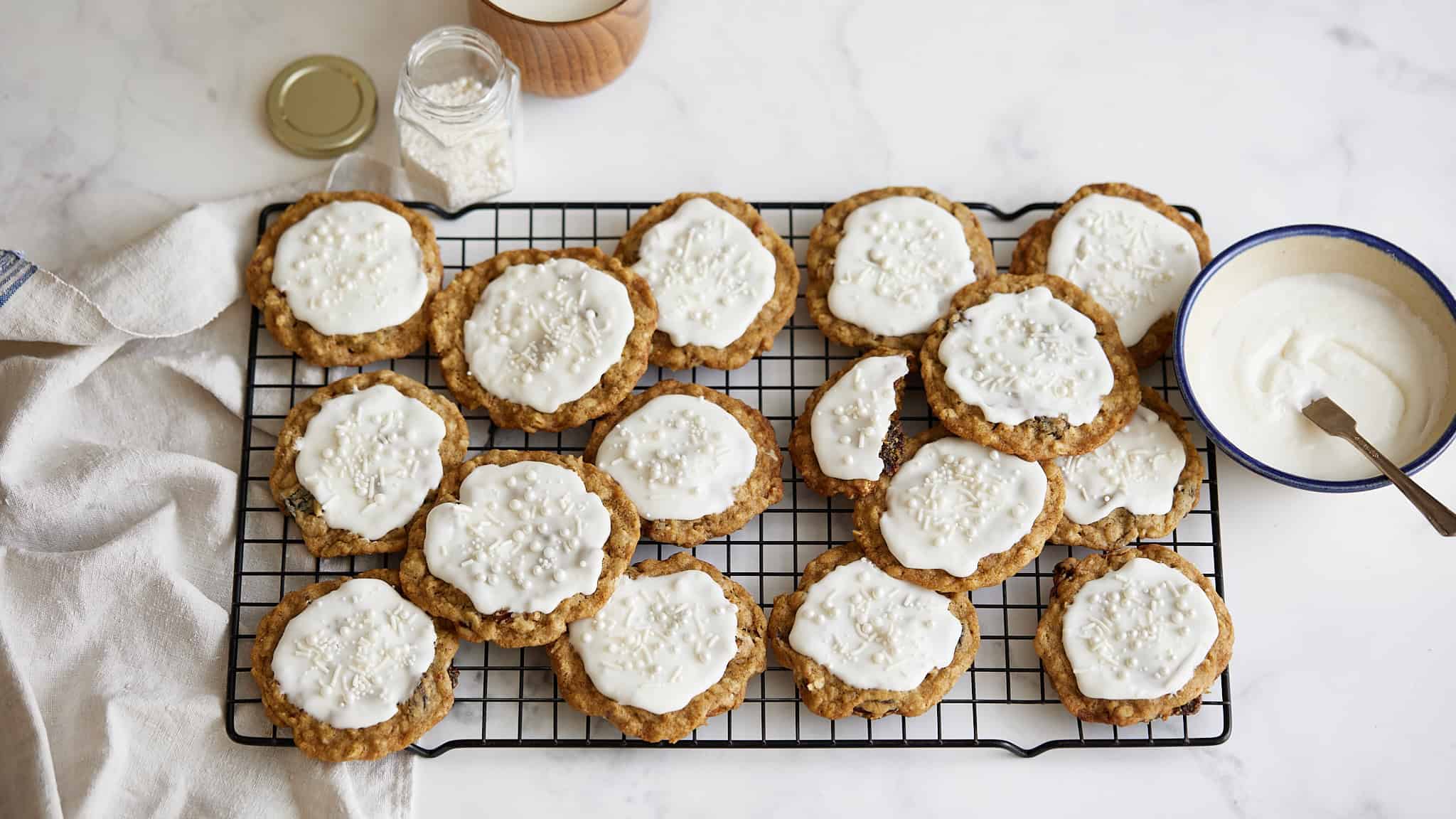 Soft cookies topped with white icing and sprinkles cooling on a wire rack with a bowl of icing and ingredients nearby.