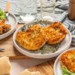 Crispy breaded chicken breasts served with fresh herbs on a white plate, surrounded by side salads and glass water cups, on a rustic wooden table with a striped cloth.