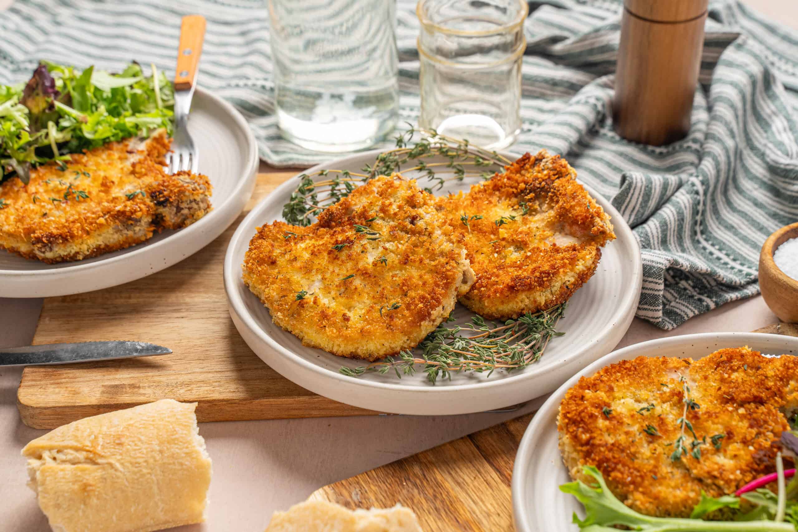 Crispy breaded chicken breasts served with fresh herbs on a white plate, surrounded by side salads and glass water cups, on a rustic wooden table with a striped cloth.