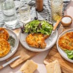Crispy breaded chicken cutlets served with fresh mixed salads on white plates, surrounded by glasses, salt, pepper, and olive oil, on a rustic wooden table for a delicious and healthy meal.