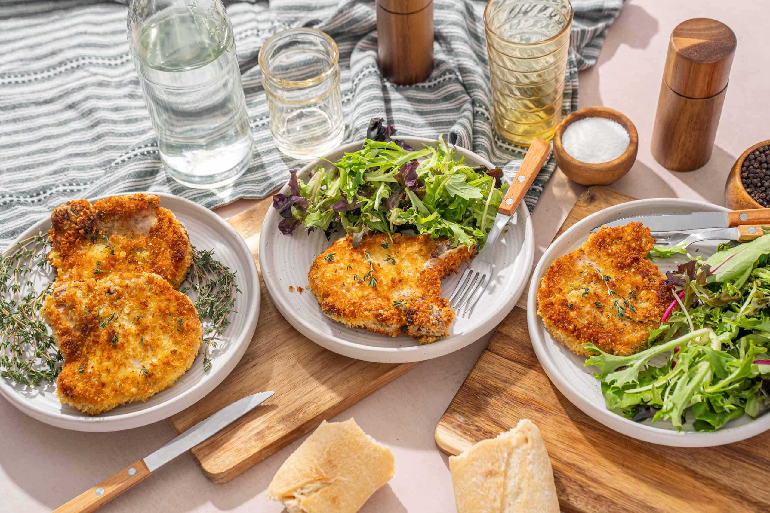 Crispy breaded chicken cutlets served with fresh mixed salads on white plates, surrounded by glasses, salt, pepper, and olive oil, on a rustic wooden table for a delicious and healthy meal.