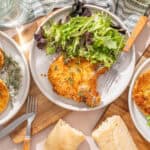 Crispy breaded chicken cutlets served with mixed green salads and fresh bread rolls on wooden platters. Perfect for a healthy, flavorful meal with herbs and a side of salt.