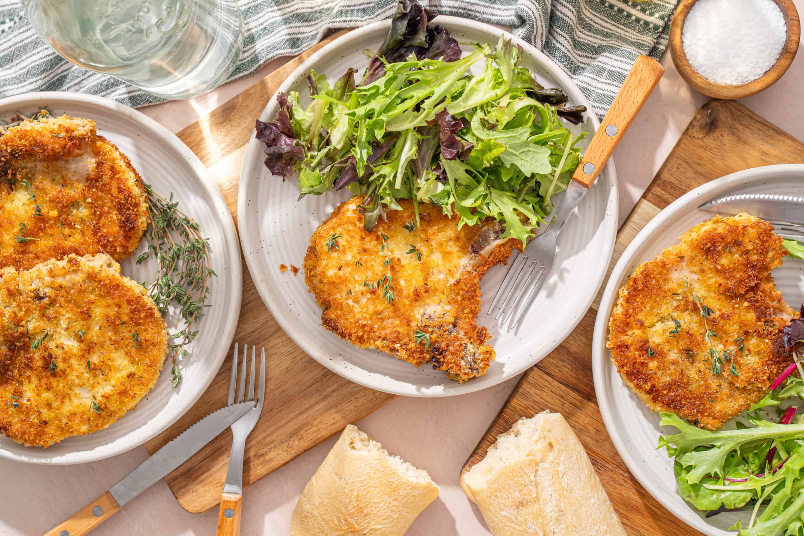 Crispy breaded chicken cutlets served with mixed green salads and fresh bread rolls on wooden platters. Perfect for a healthy, flavorful meal with herbs and a side of salt.