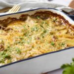 Golden baked cheesy beef and pasta casserole in a white and blue baking dish, topped with fresh chopped herbs.