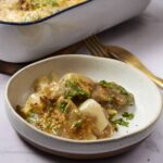 Soft chicken and vegetable stew with herbs in a white bowl, ready to serve, served alongside baked casserole dish on a wooden tray, garnished with chopped parsley, comforting homemade meal.