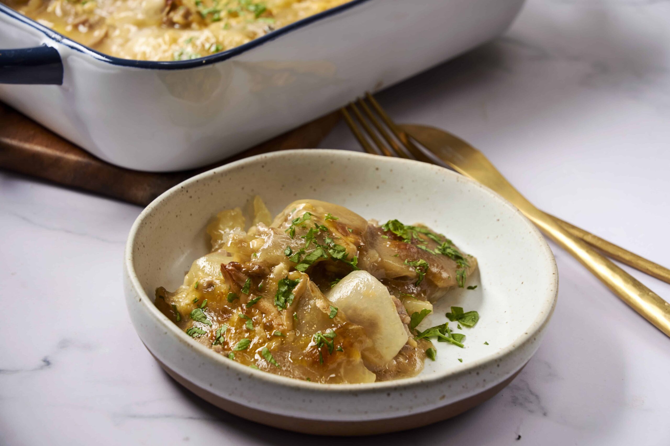 Soft chicken and vegetable stew with herbs in a white bowl, ready to serve, served alongside baked casserole dish on a wooden tray, garnished with chopped parsley, comforting homemade meal.