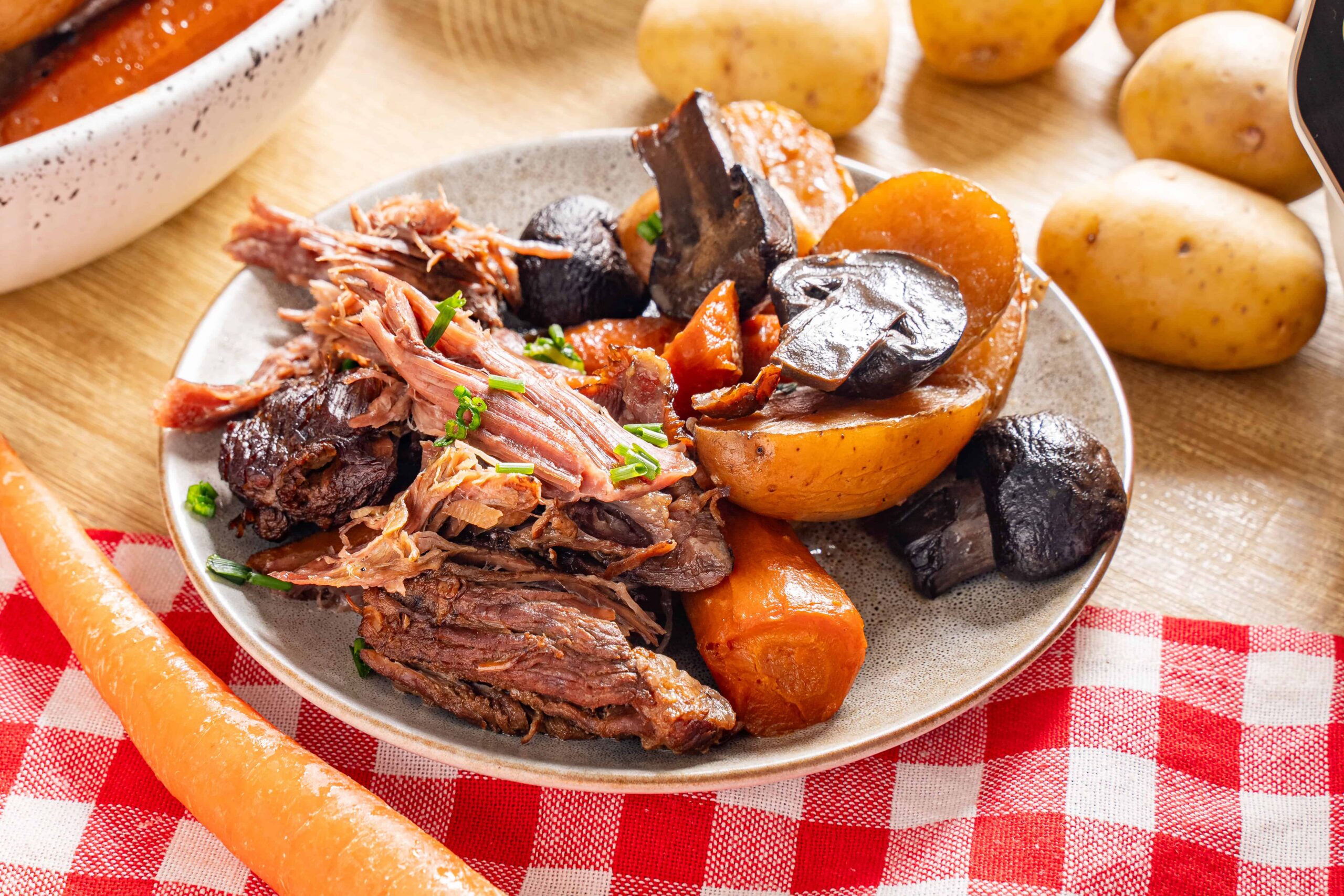Juicy shredded beef with roasted potatoes and carrots on a ceramic plate, garnished with chopped green onions, served on a wooden table with potatoes and a red checkered cloth in the background.