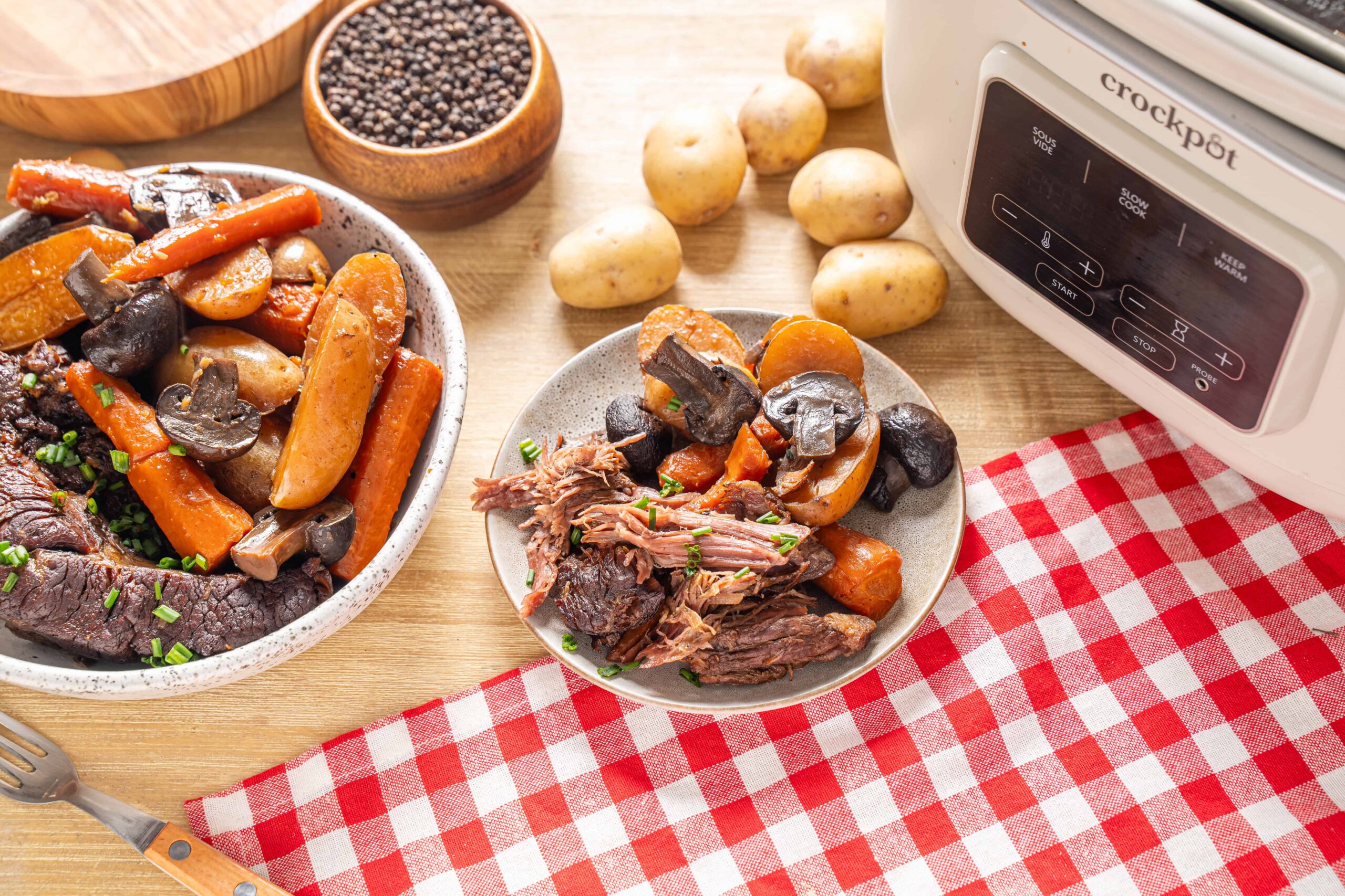 Roast beef with vegetables in a bowl, slow cooker, fresh potatoes, carrots, and mushrooms on wooden table with red-checkered cloth, ready for delicious beef stew.