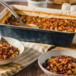 Creamy three-bean chili in a black baking dish with two bowls of prepared chili on a rustic table. Perfect for hearty, plant-based meals or comfort food recipes.