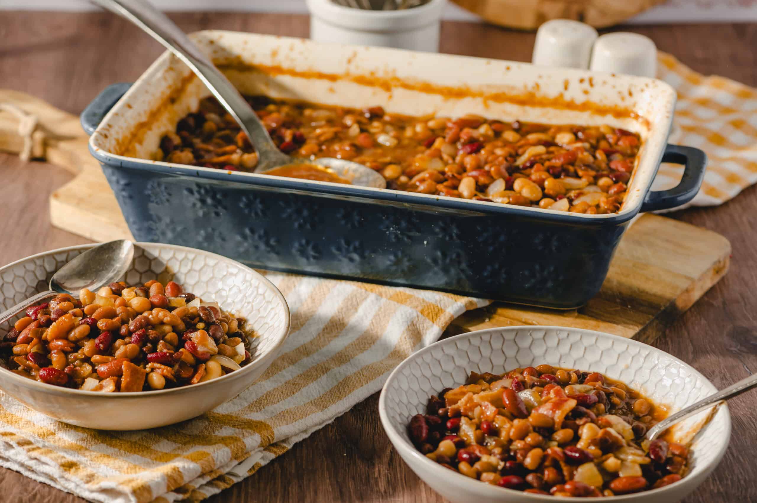 Creamy three-bean chili in a black baking dish with two bowls of prepared chili on a rustic table. Perfect for hearty, plant-based meals or comfort food recipes.