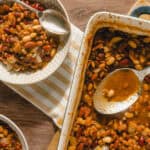 Hearty baked bean casserole served in a white dish with a spoon, featuring mixed beans, vegetables, and savory sauce, perfect for comfort food lovers.