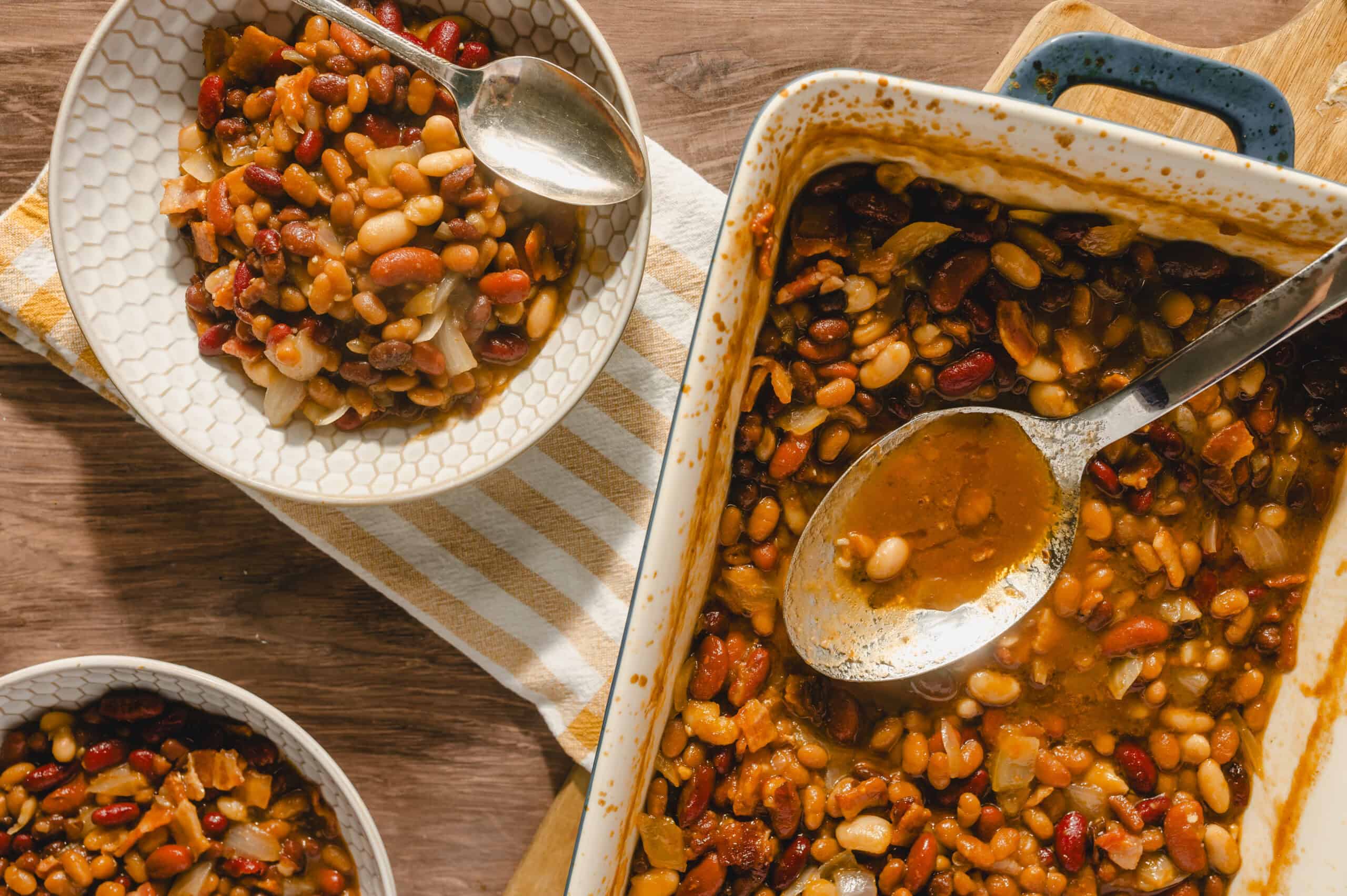 Hearty baked bean casserole served in a white dish with a spoon, featuring mixed beans, vegetables, and savory sauce, perfect for comfort food lovers.