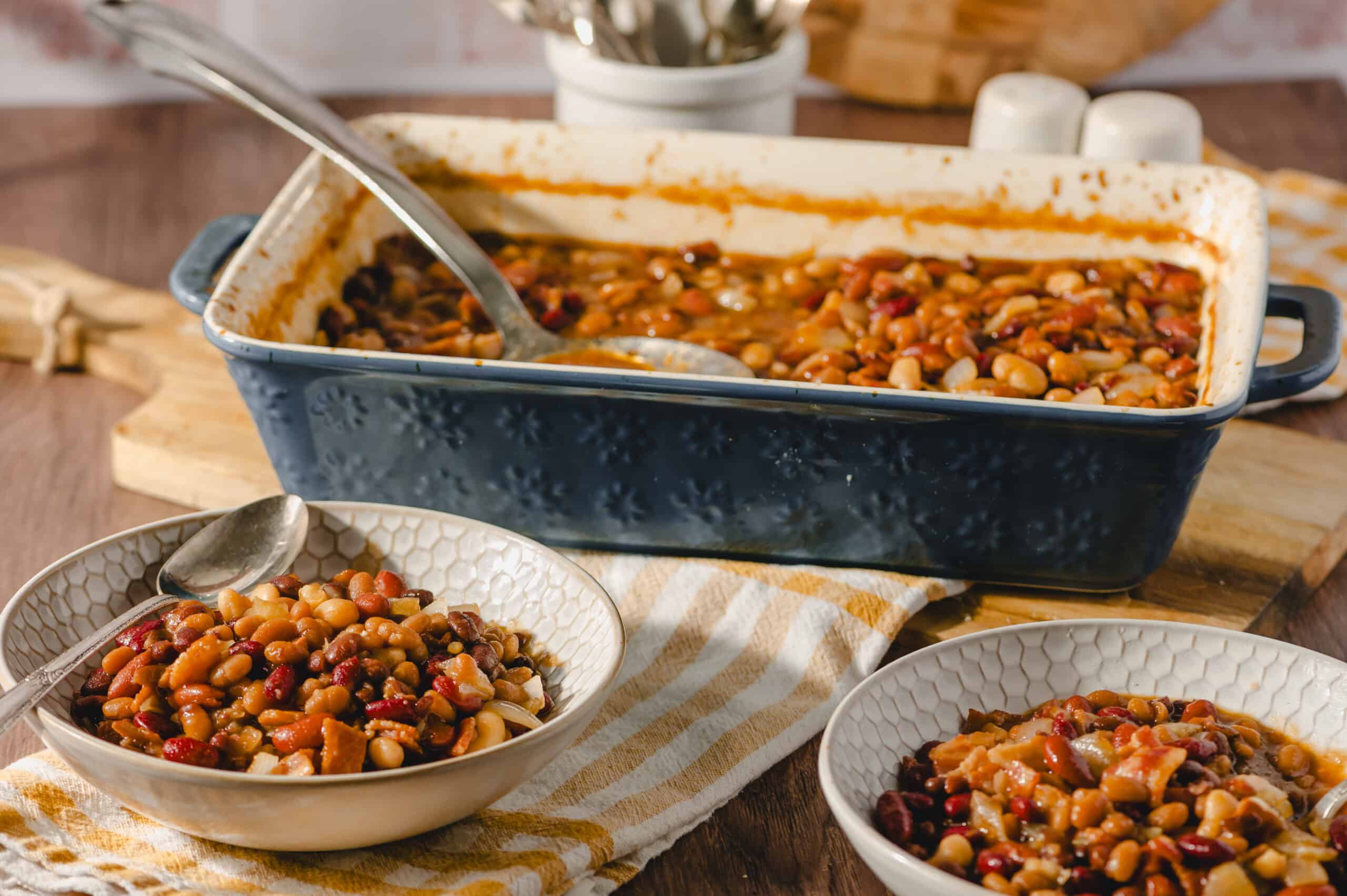 Hearty baked bean casserole served in a vintage baking dish with two bowls of the cooked beans on a rustic table.