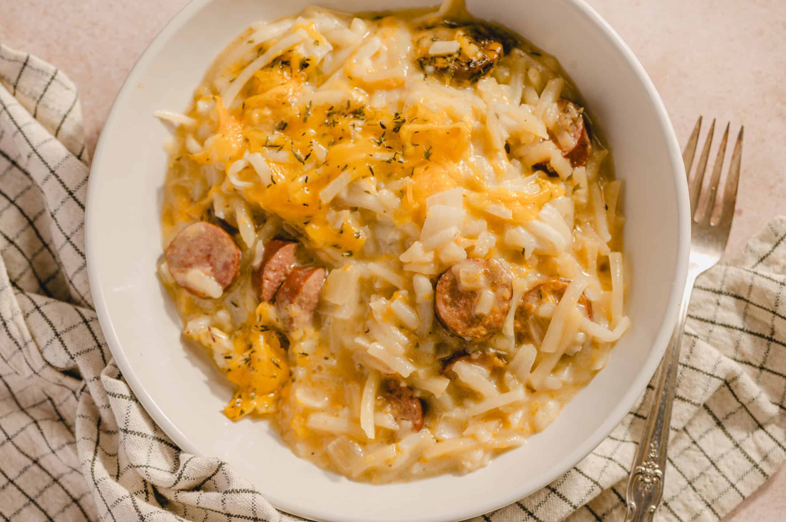 Creamy sausage and cheese potato hash casserole, with shredded cheddar and melted cheese, served on a white plate. Perfect comfort food for breakfast or brunch with sausage slices.