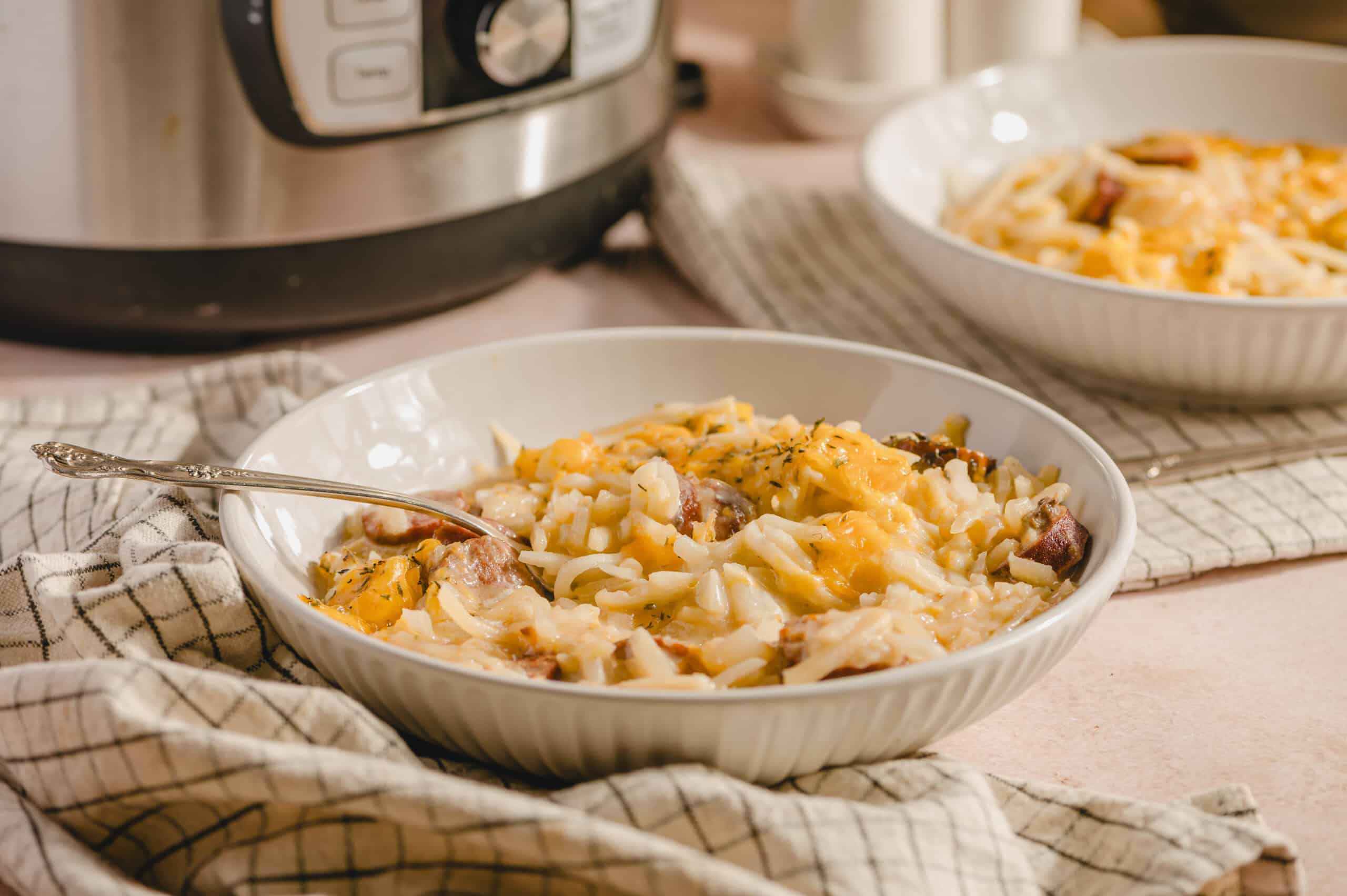 Creamy chicken and cheese casserole in a white bowl, served on a checkered cloth with a fork, featuring melted cheese, chicken, and herbs, with a slow cooker and additional casserole dish in the background.