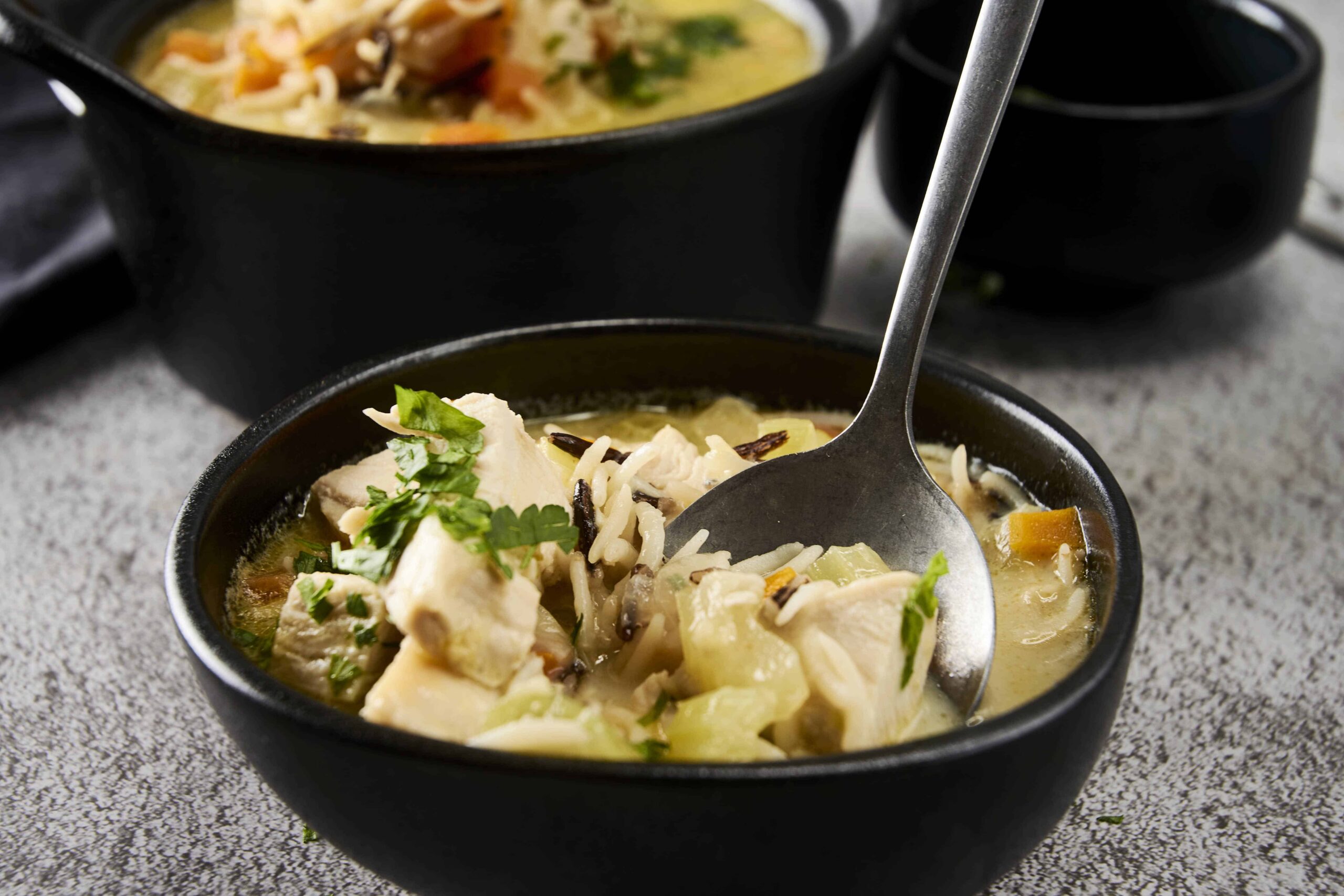 Comforting chicken and rice soup in a black bowl with fresh herbs and vegetables, served with black bowls and a spoon. Ideal for cozy home-cooked meal recipes and healthy comfort food ideas.