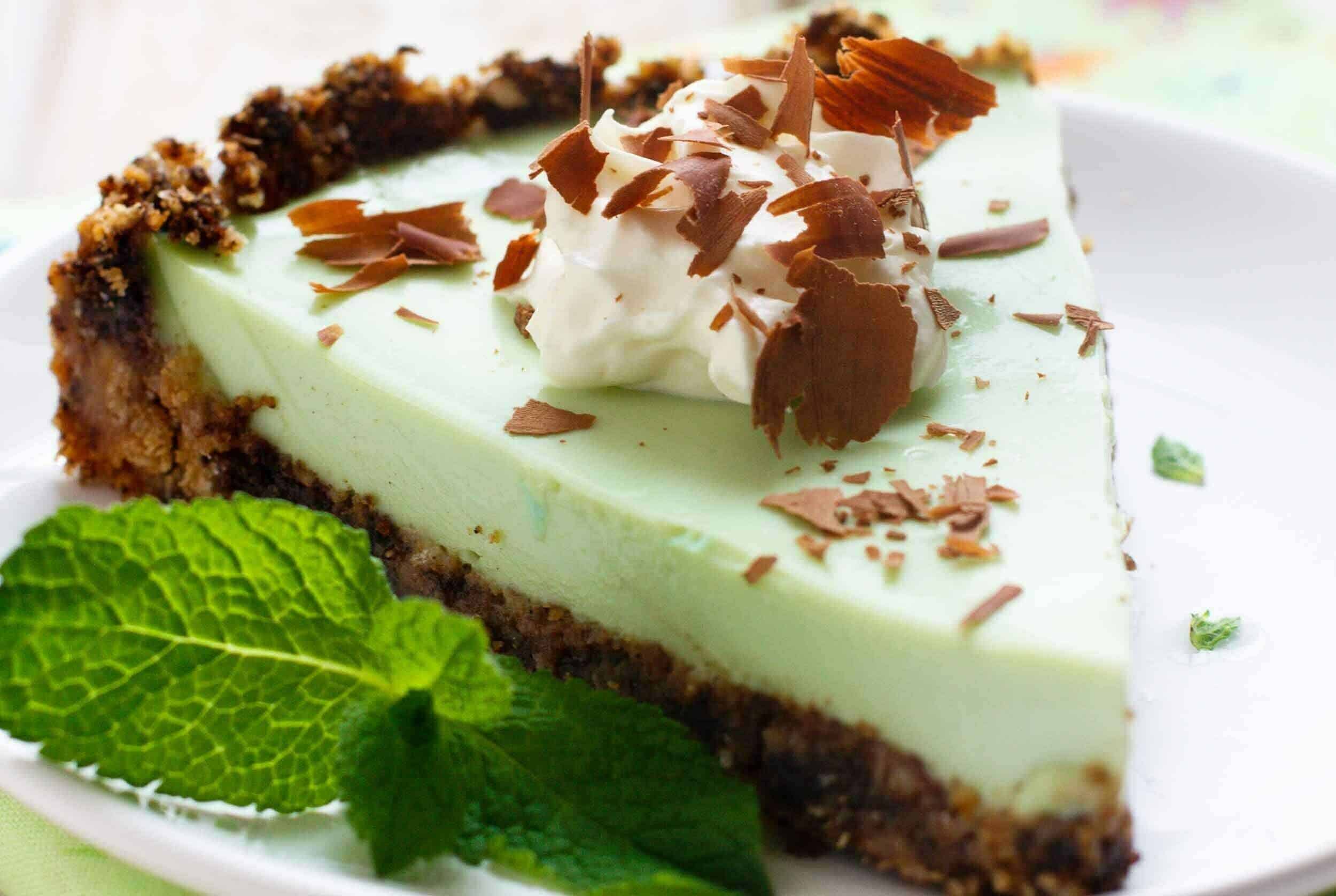 Creamy mint cheesecake with chocolate shavings and a graham cracker crust, garnished with fresh mint leaves on a white plate. Perfect for dessert lovers seeking a refreshing and indulgent treat.